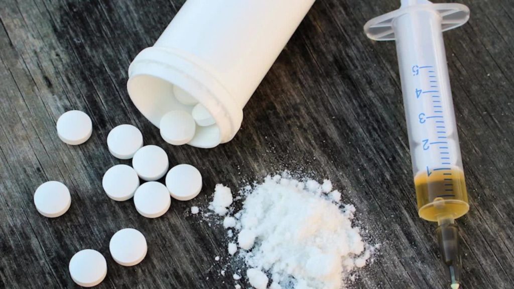 Wood table with pills, white powder fentanyl, and full syringe laying on it