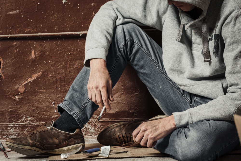 Hunched over person wearing hoodie covering face with a cigarette, staring down at needle, spoon, heroin on cardboard in abandoned building 