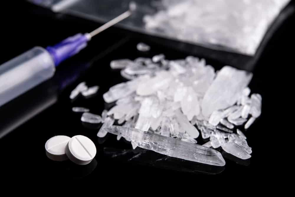 Pile of large crystal meth shards near two small pills with a syringe in the background