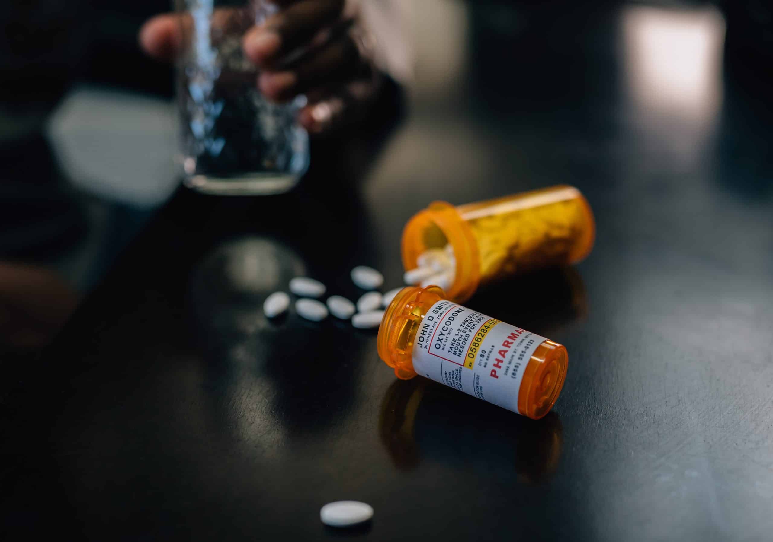 Two prescription oxycodone bottles tipped over in focus, man in need of prescription drug abuse treatment
holding glass of water in the background