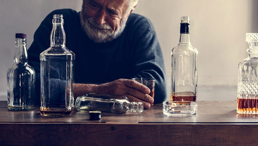 An older gentleman experiencing symptoms of alcoholism cirrhosis sits at a table with liquor bottles and a drink in his hand. 