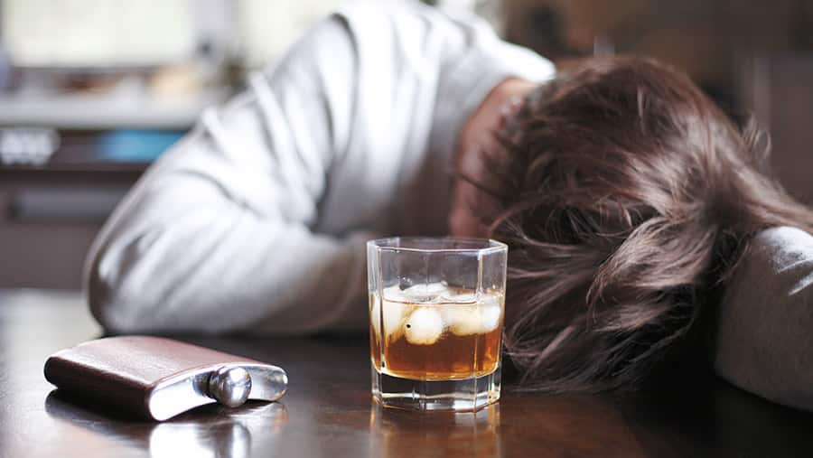 A person is slumped over a table next to a flask and glass of liquor after heavy alcohol consumption.  