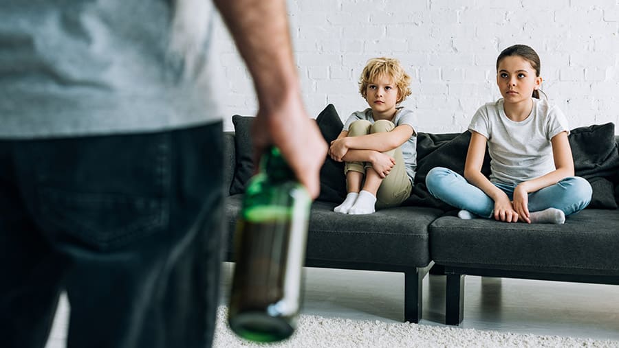 Two young school-aged children are wondering how to help and not enable their alcoholic parent who is holding a bottle of alcohol in front of them. 