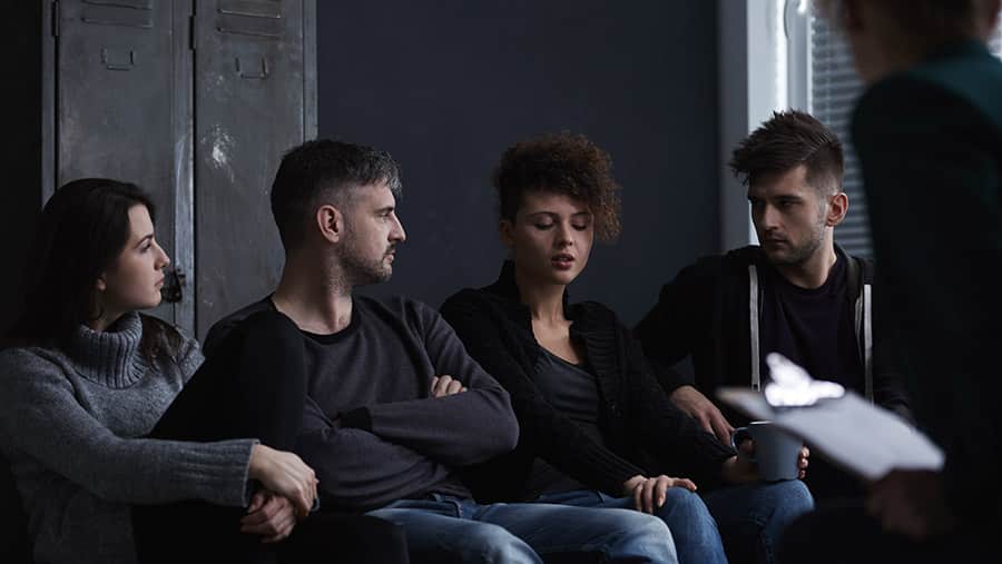 In a dark room, a group of four young people are sitting in a circle participating in dual diagnosis treatment and therapy. 