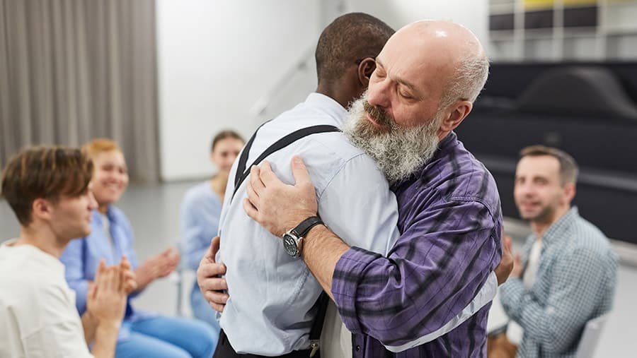 Two men are embracing in a hug surrounded by peers at a meeting addressing early sobriety and how to stay sober after rehab.