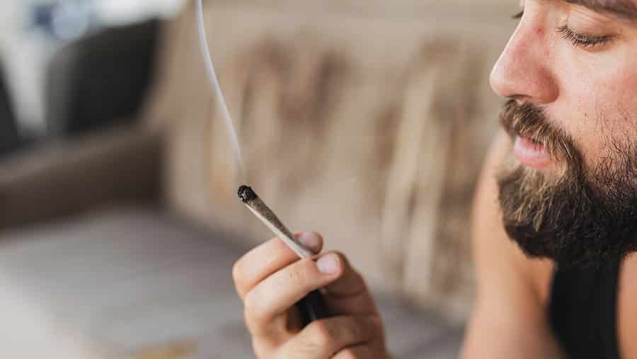 A bearded male is holding a lit blunt while contemplating quitting marijuana and experiencing the symptoms of withdrawal. 