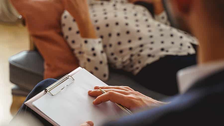 A person participating in CBT and DBT therapy is talking with a professional therapist who is holding a clipboard with a paper and pen. 