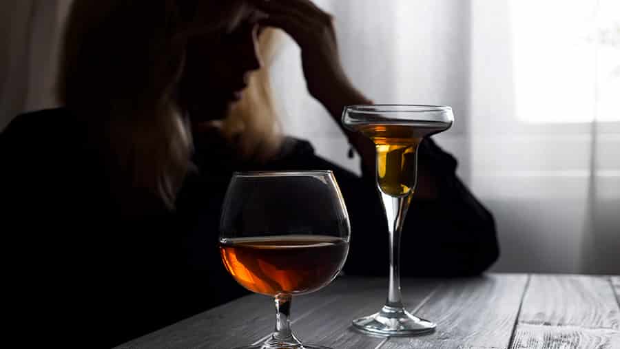 A distressed looking female who suffers from alcoholism, withdrawal, and delirium tremens sits at a table with two glasses of alcohol. 