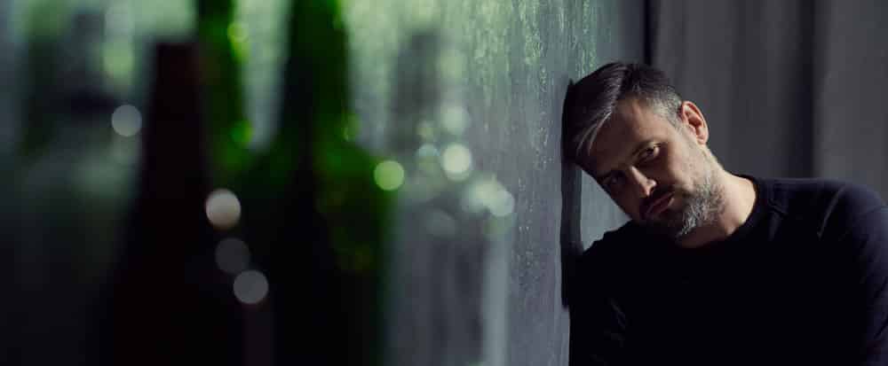 Man in need of drug detox with grey hair and beard laying his head against a dark wall with many alcohol bottles out of focus