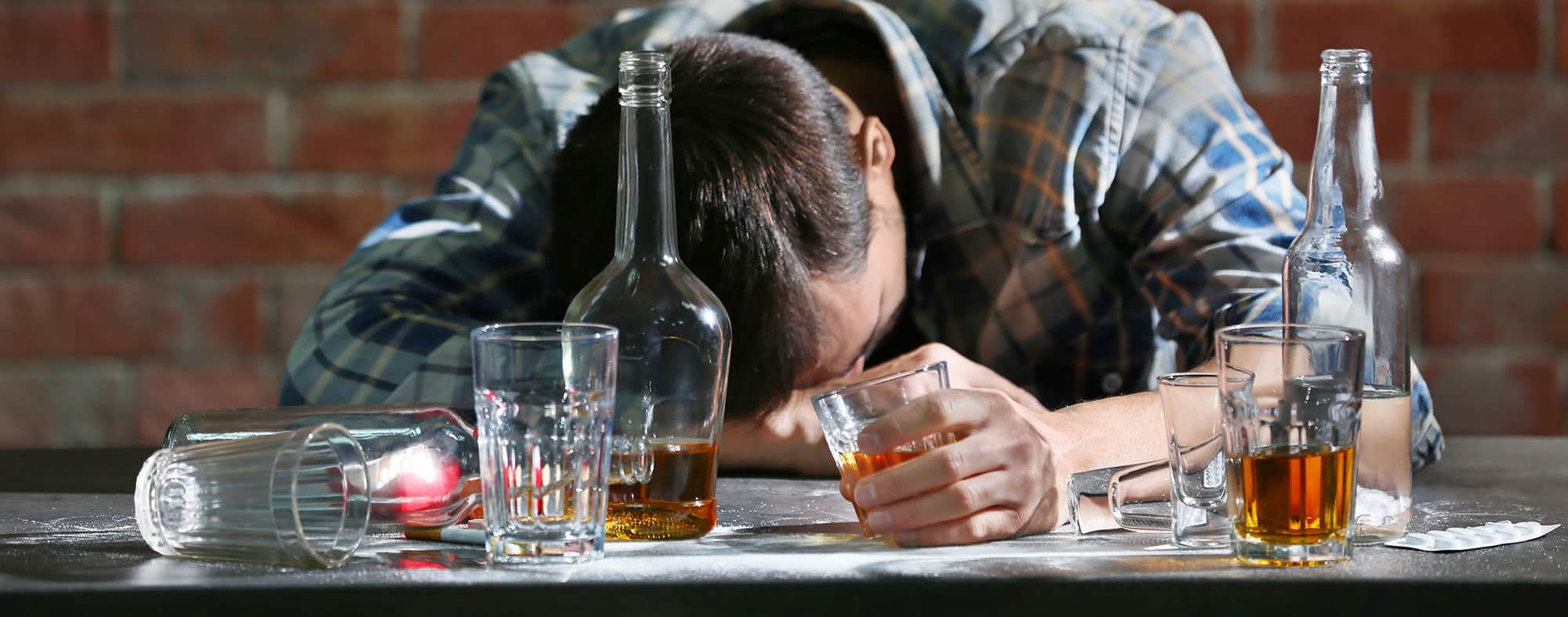 Man in plaid shirt hunched over many whiskey glasses, passed out drunk