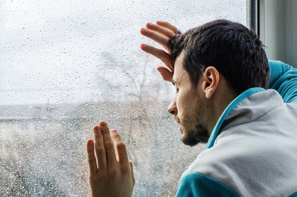Younger man with beard, in need of drug and alcohol rehab,  leaning his head against a rainy window 