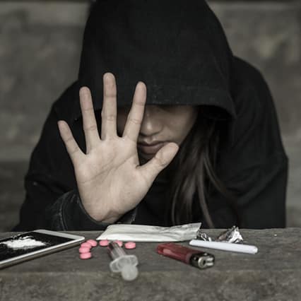 A girl in a hoodie covering her eyes puts her hand up in a stop motion with illegal drugs below it on a the table
