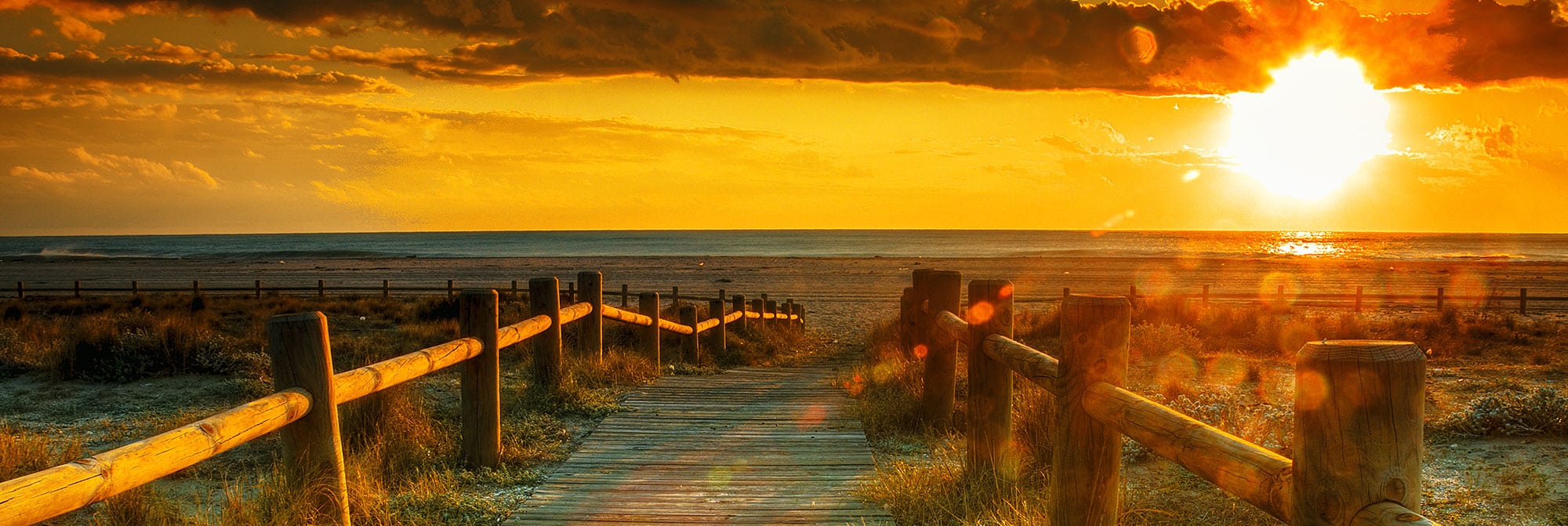 Wood fenced boardwalk going towards sun shining brightly under clouds and setting on the ocean in the horizon, 