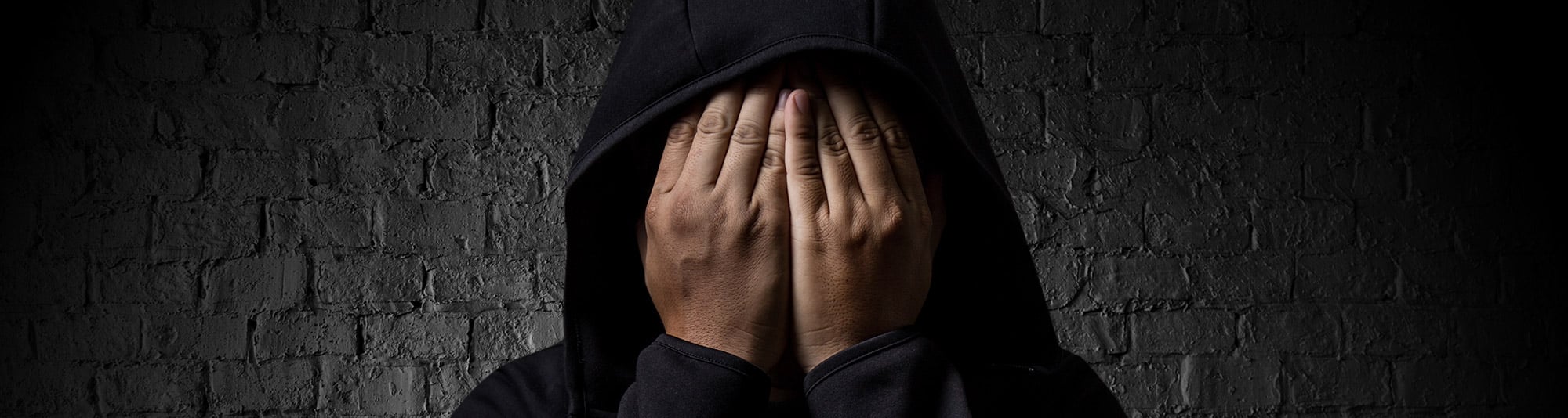 Girl in dark room with grey bricks, covers face with hands while wearing a hoodie