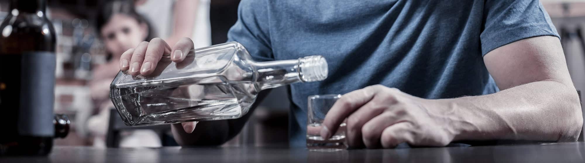 Close up of man pouring alcohol into a shot glass with his daughter behind him watching from afar