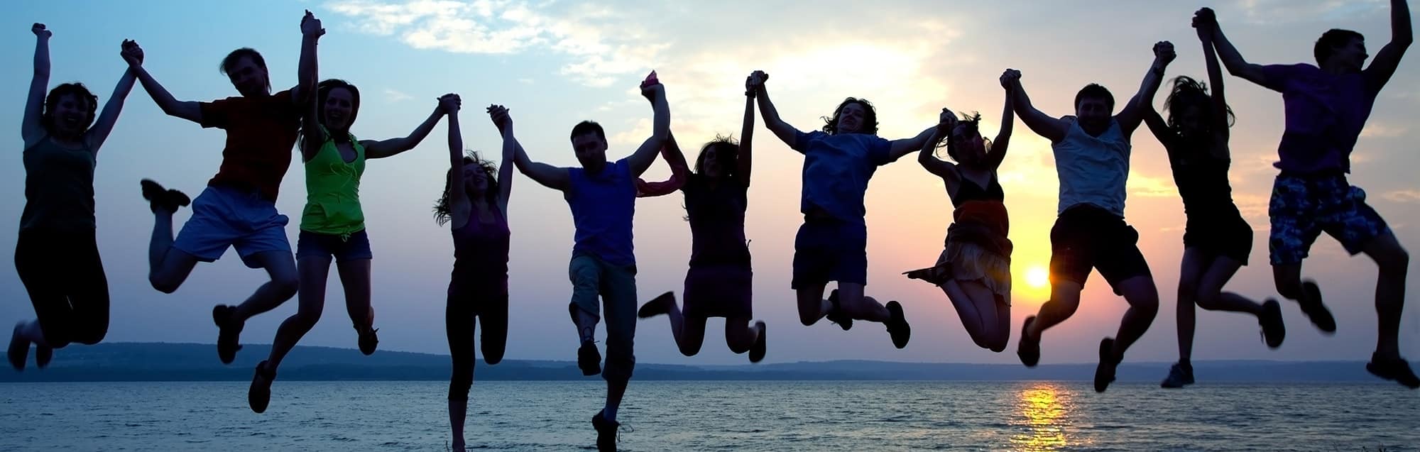 Large group of young adults holding hands and jumping in the air with the ocean behind them and the sun setting