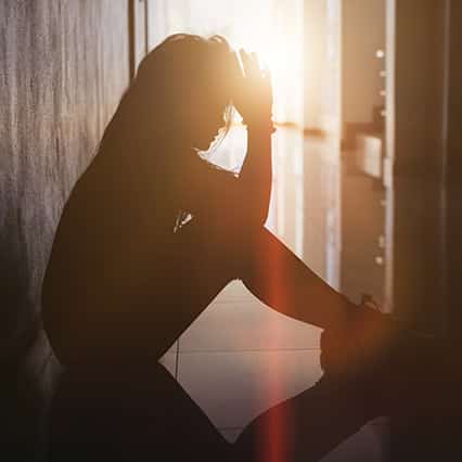 dark profile of young woman sitting on the floor with head in her hands and sun coming through
