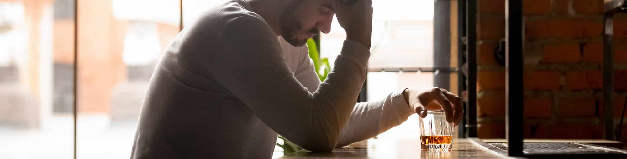 Man in need of drug and alcohol rehab, sitting at a bar, looking down at glass of alcohol