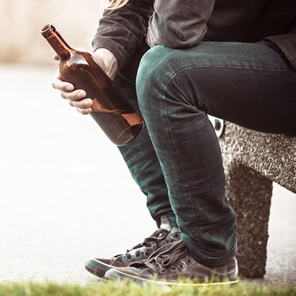 bottom half of man in hoodie and jeans sitting on a bench with bottle of alcohol in hand
