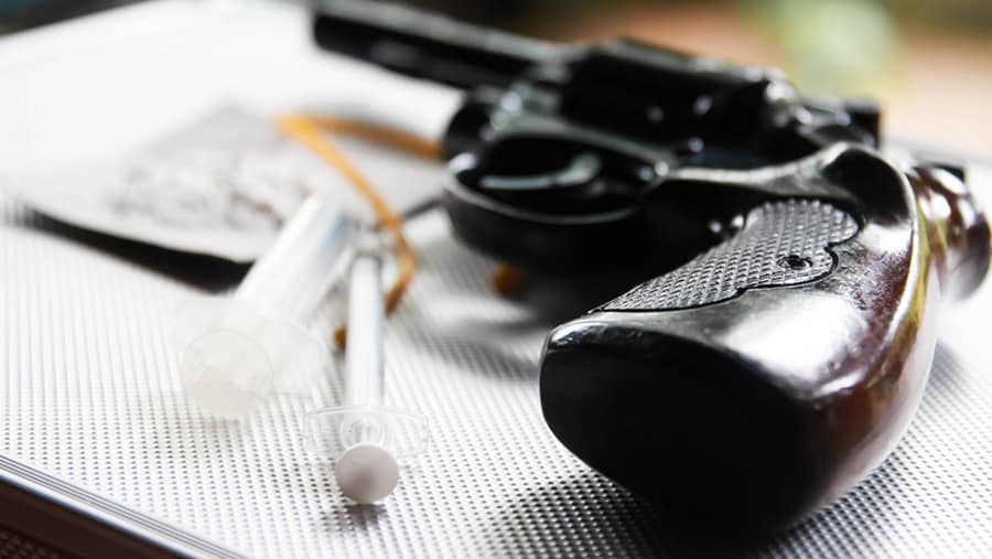 A handgun and needle syringes are on a silver metal table. 