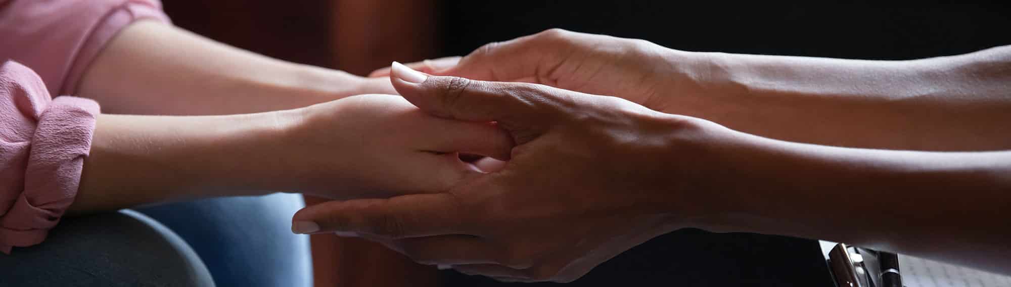 Close up of counselor's hands holding another person's hands with their knees almost touching. Clipboard on counselor's lap