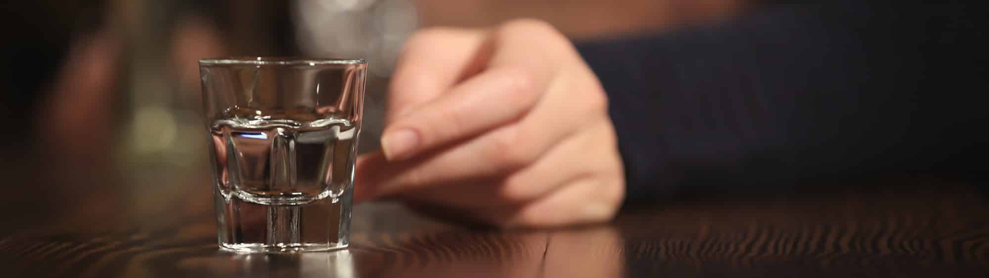 Empty shot glass in focus with hand out of focus reaching for it