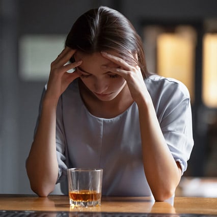 Girl looking down at glass of whiskey, head in hands, contemplating if she should drink it