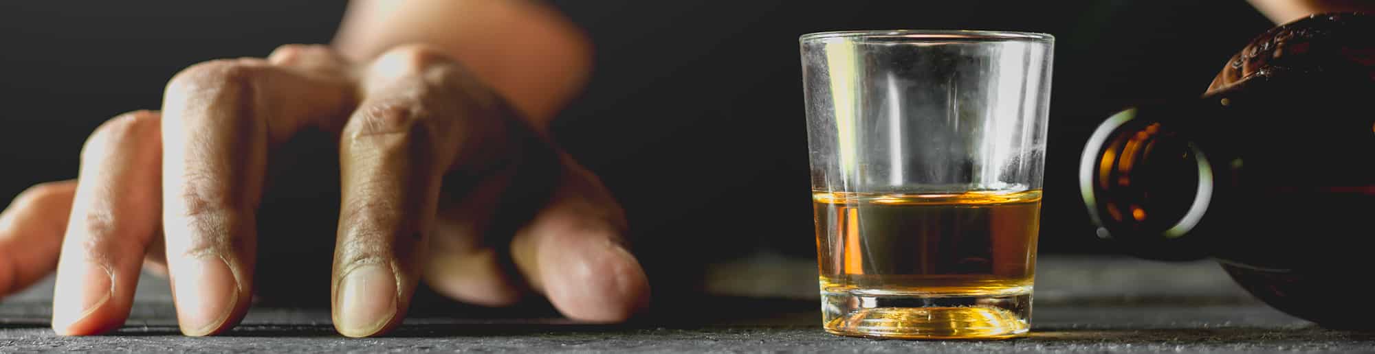Hand rested near shot glass of whiskey with bottle lying next to it