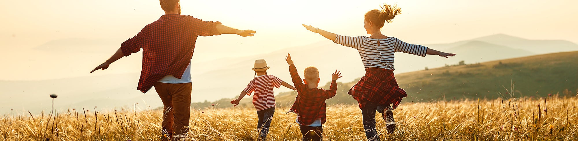 Family of mom, dad, and two kids running in a wheat field towards a lake with the sun shining brightly