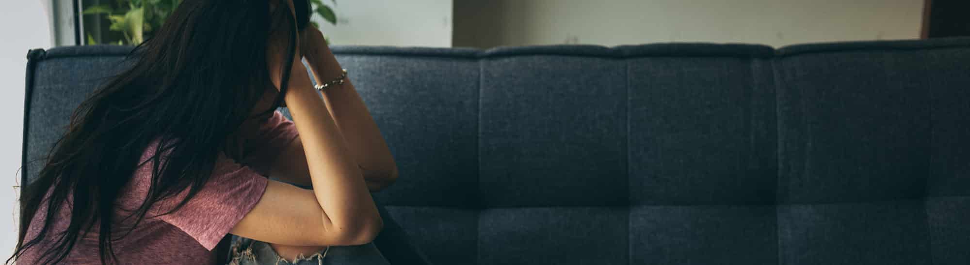 Side profile of woman with long dark hair, sitting on couch with head in hands