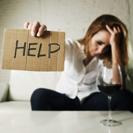 Woman in need of alcohol rehab in ohio, with head down in hand and wine glass next to her on a table, holding up sign that says "HELP"