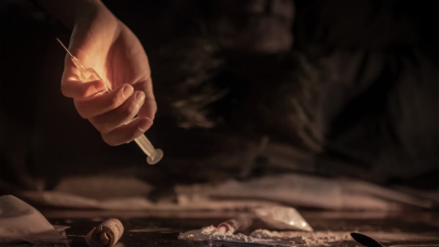 A hand of a person experiencing a drug overdose holding a needle above a dark table with rolled-up bills, small plastic bags and white powder. 