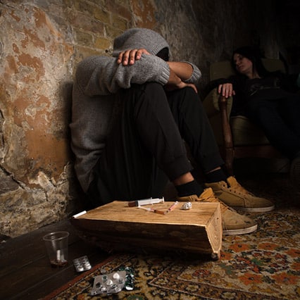 Guy in need of rehab, sitting against the wall with his head down and knees against chest, syringes and drugs next to him on a wood table