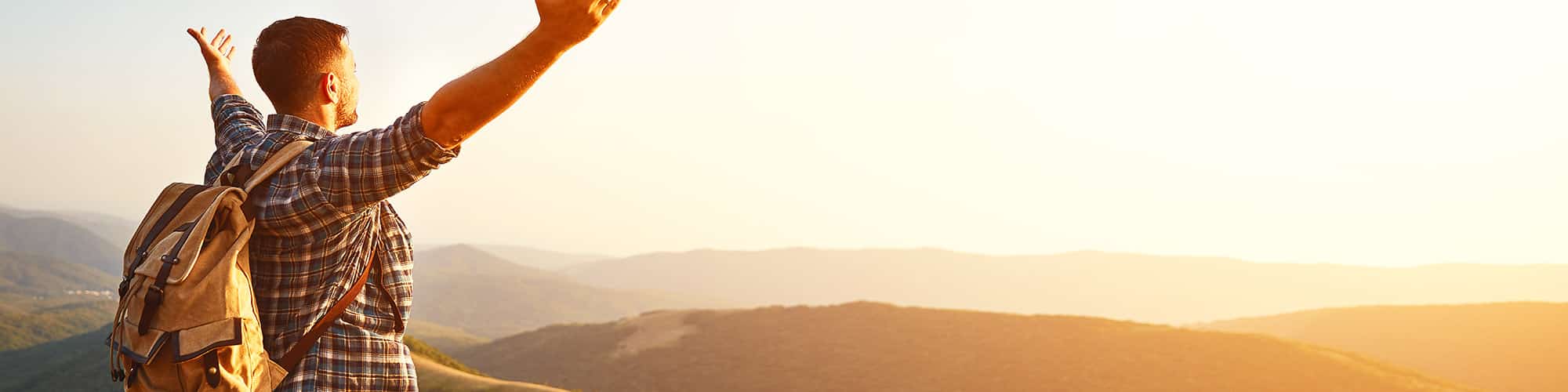 Young man at the top of mountain with a hiking backpack on, his arms are up looking towards the sun and along mountain tops