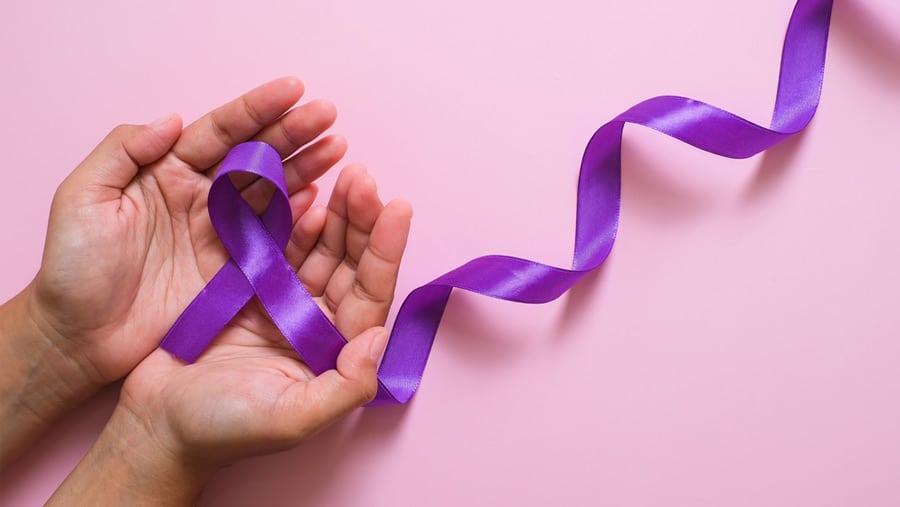 A person’s hands holding a purple ribbon representing International Overdose Awareness Day on a pink background. 