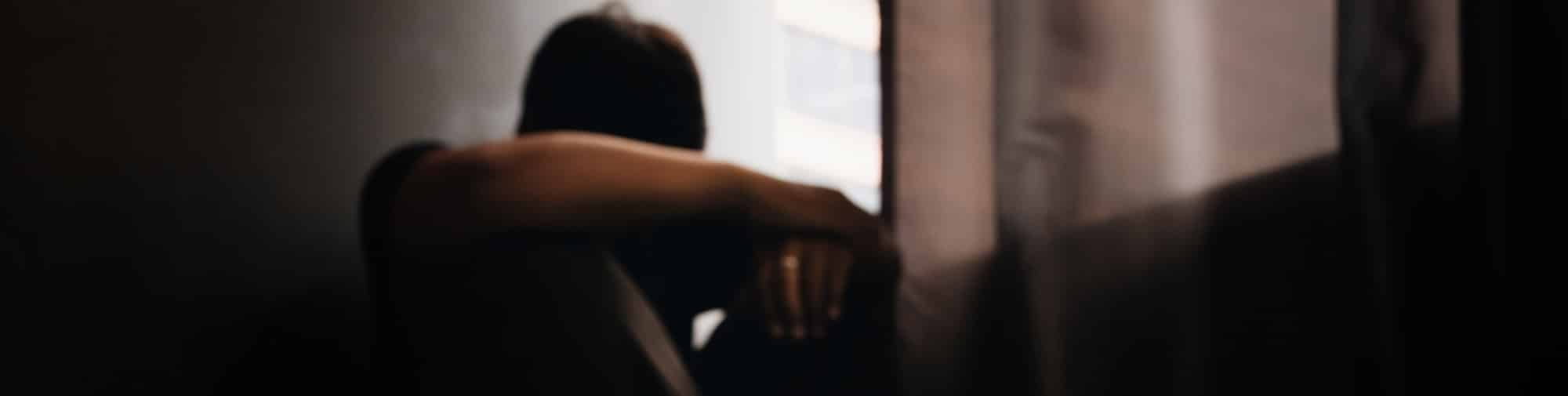 Blurry, out of focus, man sits against a wall in a dark room with his head down and arm covering his face