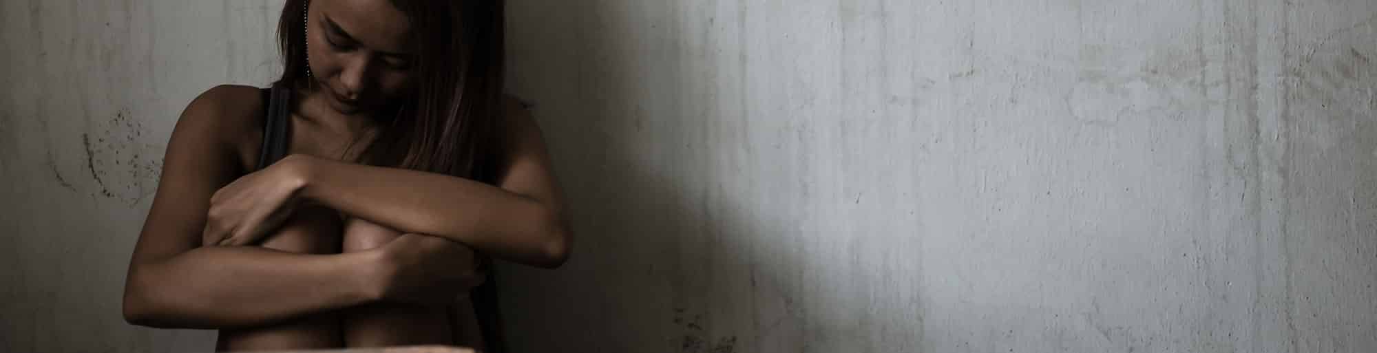 Girl sitting in scrunched up position, holding her knees close to her chest in a dark room against a grey wall