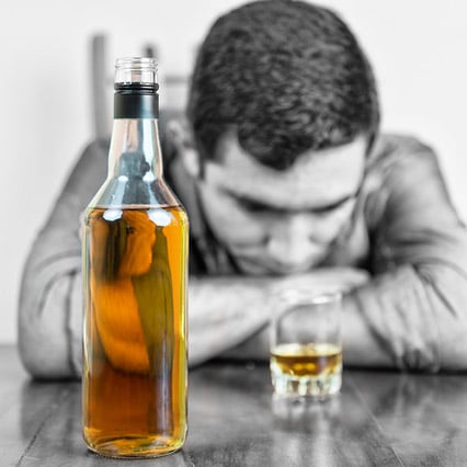 Black and white man resting on arms staring at gold glass of whiskey with a gold whiskey bottle in view