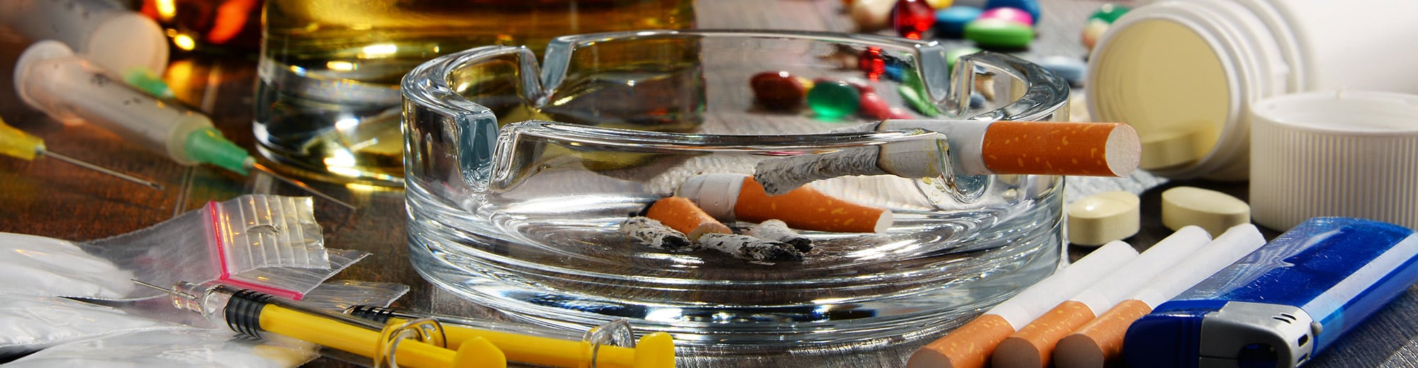 Ashtray with many cigarette butts in it and one lit cigarette resting on edge with cigs, lighters, syringes, pills, pill bottles in the background