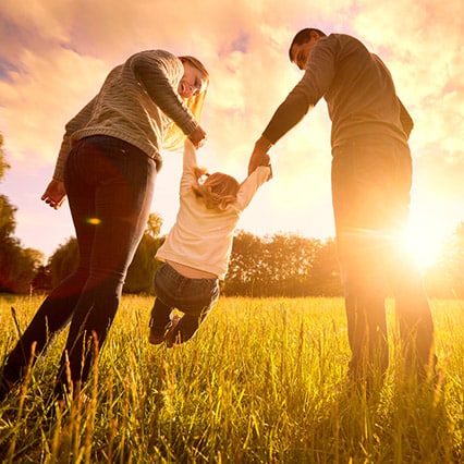 Parents, each holding a hand of little girl swinging her in a green field with trees surrounding and the sun shining
