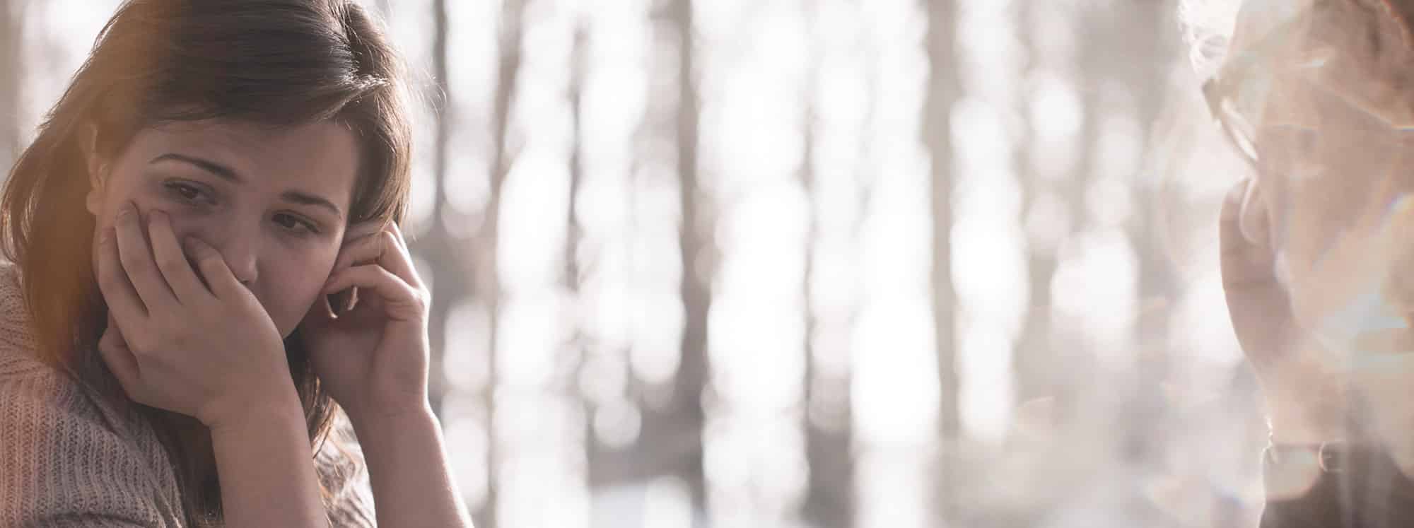 young girl looking away sad with hand over her mouth with older woman staring at her, surrounded by trees in the background 