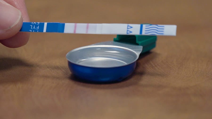 A blue and white fentanyl test strip shows two pink lines next to a small round bowl of clear liquid containing the liquid fentanyl. 