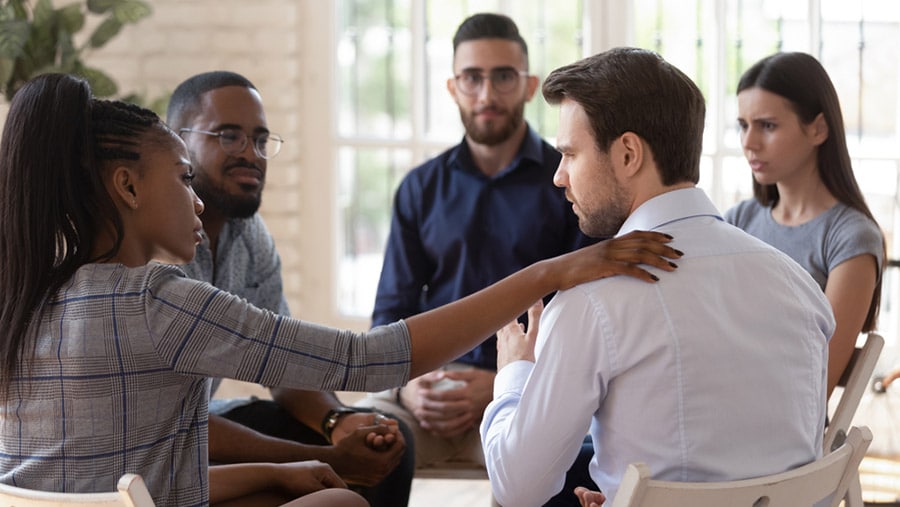 A group of young adults in an addiction recovery support group meeting, sitting in a circle looking concerned over a young man. 