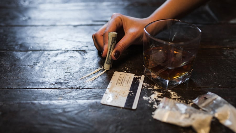 A table with a glass of alcohol next to a hand holding a rolled-up dollar bill above two lines of cocaine surrounded by a card and small plastic bags covered in white powder. 