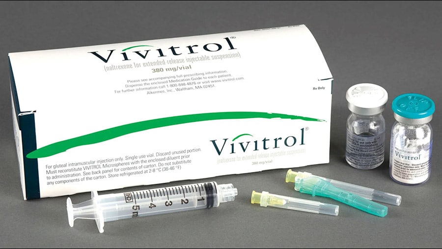 A large box packaging of Vivitrol surrounded by a syringe, and two small glass vials containing Vivitrol medication.