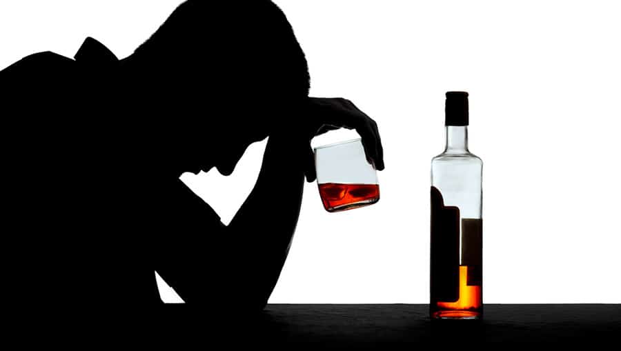 Man in dark shadow taking the first step toward treatment for alcohol addiction holding a glass of liquor next to an opened liquor bottle. 