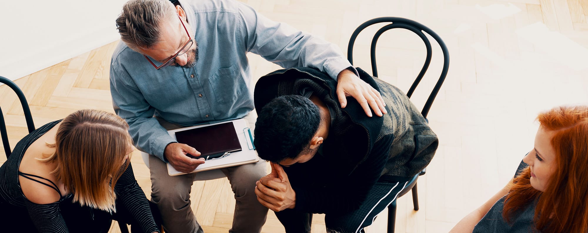 Four people sitting in a semi-circle, one man sitting with his head down resting on his arms while another older man consoles him, with his arm resting on his shoulder