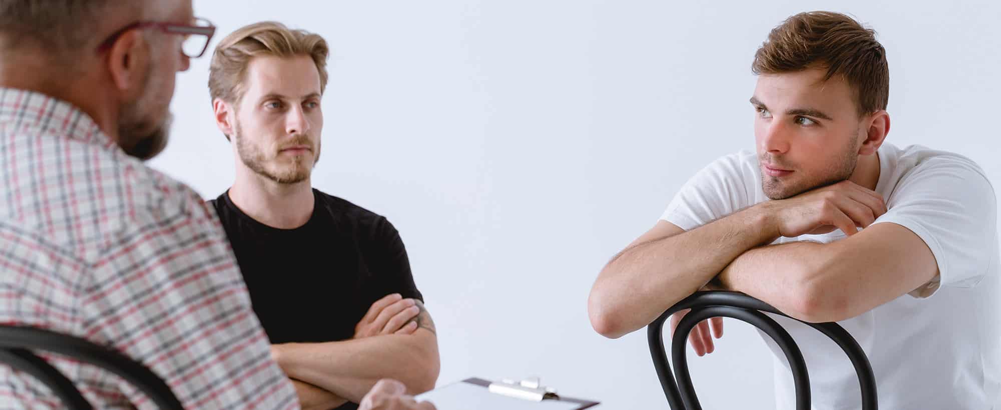 Three men sitting faced towards each other in a brightly lit room during group therapy in a men's rehab