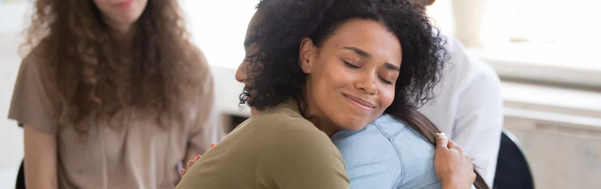 Two women embracing in a supportive hug during therapy with two woman in the background watching in a brightly lit room