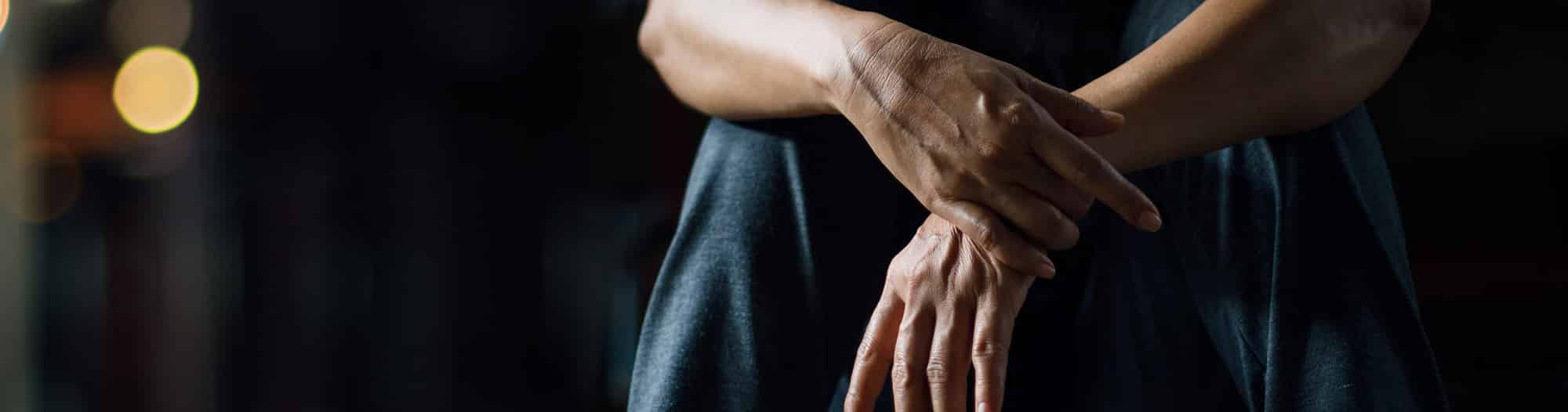 Close up of two hands resting around waist, one is clutching the other arm's wrist with a dark background and lighting